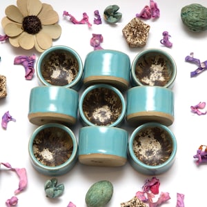 May include: Nine small, cylindrical ceramic bowls with a light blue exterior and a speckled brown interior. The bowls are arranged in a grid pattern on a white surface, surrounded by dried flowers and decorative elements.