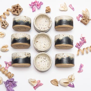 May include: An overhead view of nine small ceramic pots and bowls. The pots have a speckled white and black glaze over a dark band, with a natural brown base. The bowls are speckled white. The arrangement is decorated with dried flowers and decorative elements.