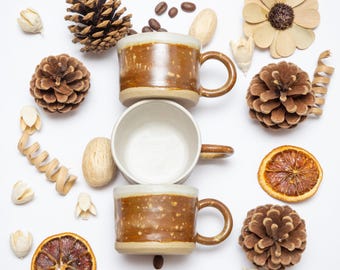 May include: Overhead shot of two brown and cream-coloured ceramic mugs with handles, stacked on top of each other. The mugs are surrounded by pine cones, dried orange slices, coffee beans, and decorative elements on a white background.