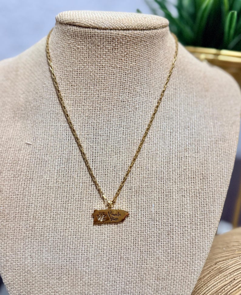May include: Gold-toned necklace featuring a pendant shaped like the island of Puerto Rico. The pendant has the words "Puerto Rico" engraved on it. The necklace is displayed on a mannequin.