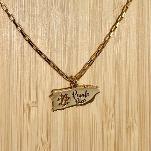 May include: Gold-toned necklace featuring a pendant in the shape of Puerto Rico. The pendant has the words "Puerto Rico" and a small design. The necklace is displayed against a wooden background.