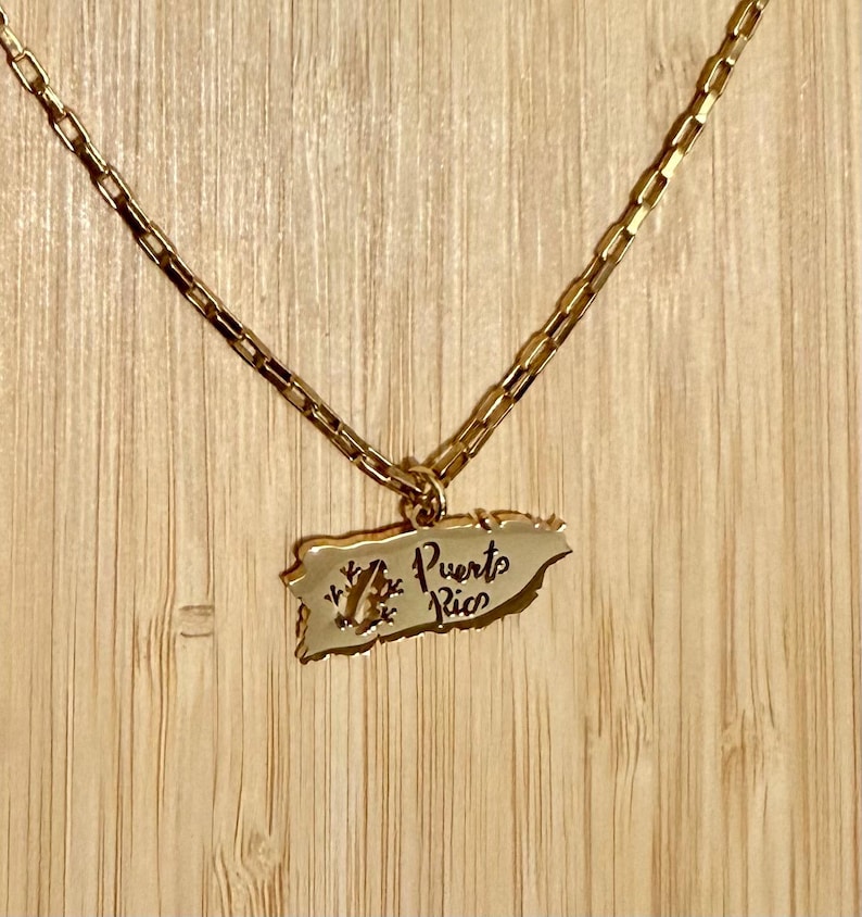 May include: Gold-toned necklace featuring a pendant in the shape of Puerto Rico. The pendant has the words "Puerto Rico" and a small design. The necklace is displayed against a wooden background.