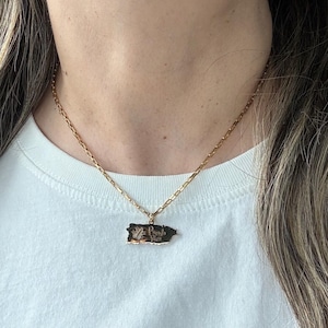May include: Gold-toned necklace with a rectangular pendant in the shape of Puerto Rico. The pendant has a detailed design and text. The necklace is worn over a white t-shirt.