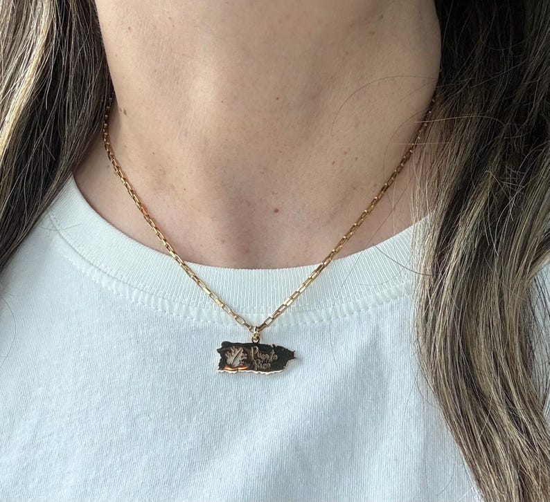 May include: Gold-toned necklace with a rectangular pendant in the shape of Puerto Rico. The pendant has a detailed design and text. The necklace is worn over a white t-shirt.