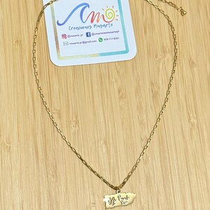 May include: A gold-colored necklace with a rectangular chain and a Puerto Rico pendant. The pendant features the outline of the island with the words "Vive Puerto Rico" in script. A business card with the text "Creaciones Musarte" is in the background.