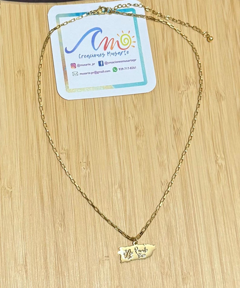 May include: A gold-colored necklace with a rectangular chain and a Puerto Rico pendant. The pendant features the outline of the island with the words "Vive Puerto Rico" in script. A business card with the text "Creaciones Musarte" is in the background.