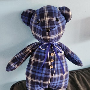 May include: A blue and white plaid teddy bear with black button eyes and a blue ribbon tied around its neck. The bear has a button closure down the front.