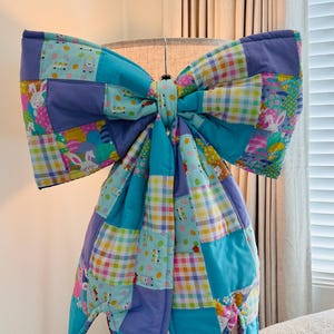 May include: A large, colorful quilted bow in shades of blue, purple, and pastel patterns. The bow is made of patchwork squares with Easter-themed designs, including bunnies and eggs. It is displayed on a lamp.