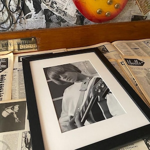 May include: A black and white photograph of a musician playing a guitar. The photo is framed in a black frame and is displayed on a table with newspapers and magazines.