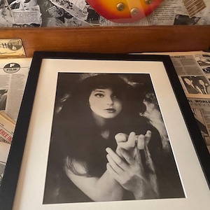 May include: A black and white framed photograph of a woman with long dark hair. She is wearing a simple top and has her hands clasped together. The photo is set against a white background.