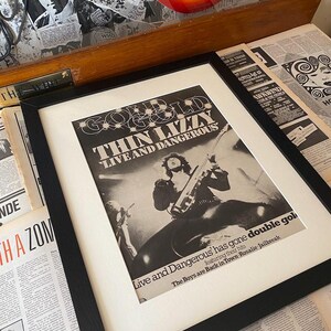 May include: A framed black and white concert poster for Thin Lizzy's "Live and Dangerous" tour. The poster features a photo of the band performing on stage with the text "Gold" and "Thin Lizzy" in large, bold letters. The poster also includes the text "Live and Dangerous" and "The Boys are Back in Town Rosalie Jailbreak".