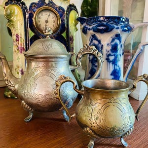 May include: An antique silver tea set with floral engravings, comprising a teapot and sugar bowl. The set has decorative handles and rests on small feet. A vintage clock and a blue and white jug are in the background.
