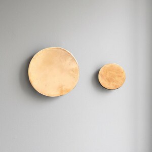 Può includere: Due decorazioni murali circolari color oro di diverse dimensioni sono montate su un muro grigio. Il cerchio più grande ha un diametro di circa 30 cm, mentre quello più piccolo misura circa 15 cm. La finitura metallica conferisce un aspetto moderno.