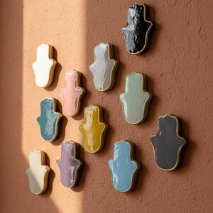 May include: A selection of ceramic Hamsa hand wall decorations in shades like white, pink, teal, yellow, and blue. Each hand features a gold-coloured trim and is fixed to a textured, reddish-brown wall. The arrangement is artistic and decorative.