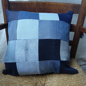 Può includere: Un cuscino in patchwork di denim blu con diverse tonalità di tessuto denim blu. Il cuscino è seduto su un sedile di sedia intrecciato.