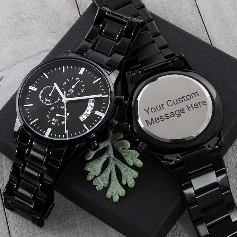 men engraved watches – a black wristwatch featuring a three-dial configuration, calendar function, and an elegant luxury pointer, is displayed on a closed black gift box. A back engraved wristwatch is also shown on the gift box that reads "your custom message here". The watches and the box are placed on a light grey background.