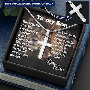 Pode incluir: Um colar com pingente de cruz prateada exibido em uma caixa de presente preta. A cruz é gravada com "Love you, Dad". A caixa tem uma imagem de leão e o texto "To my Son" e "Love Dad".