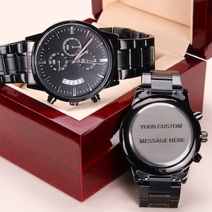 personalized watches for men – a black chronograph watch featuring a three-dial configuration, calendar function, and an elegant luxury pointer, is displayed on a closed luxury gift box. A back of a wristwatch is shown next to the gift box with an engraving that reads "your custom message here". The watches and the box are placed on a light beige background.