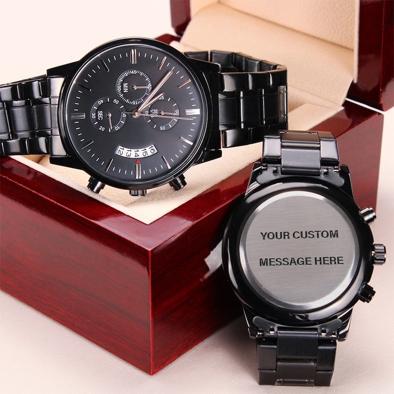 personalized watches for men – a black chronograph watch featuring a three-dial configuration, calendar function, and an elegant luxury pointer, is displayed on a closed luxury gift box. A back of a wristwatch is shown next to the gift box with an engraving that reads "your custom message here". The watches and the box are placed on a light beige background.