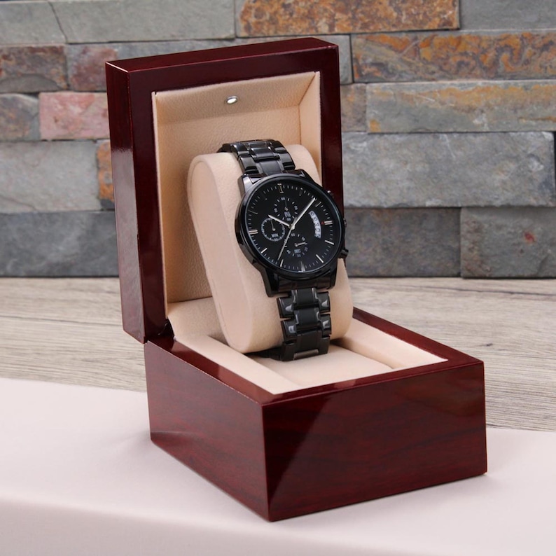 May include: A black wristwatch with a matching black band is displayed in a dark wood presentation box. The watch has a black face with white accents and three sub-dials. The box is open, revealing a beige interior.
