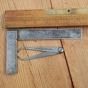 May include: A silver metal try square with a silver metal compass with the text "CHESTERMAN & SONS SHEFFIELD ENGLAND" on it. The tools are on a wooden surface.