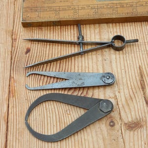 May include: Two metal calipers, one with a circular handle and the other with a hexagonal handle, are lying on a wooden surface.