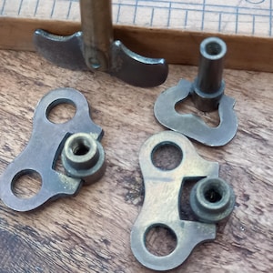 Puede incluir: Primer plano de varias piezas pequeñas de ferretería antigua de latón. Los componentes incluyen palancas, soportes con agujeros circulares y cilindros roscados. Las piezas están dispuestas sobre una superficie de madera, mostrando su pátina envejecida y diseños intrincados.