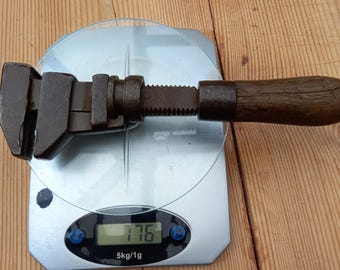 Puede incluir: Una llave inglesa antigua con mango de madera, colocada sobre una báscula digital. La llave es de color marrón oscuro y muestra signos de desgaste. La báscula muestra un peso de 176, con una medida de 5kg/1g.