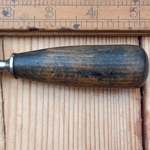 May include: Close-up of a wooden handled chisel with a metal blade. The handle is dark brown with a black stain and has a worn, aged look.