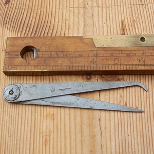 May include: A silver metal compass with a brass crown and the text "A.18  REX  QUEENSFERRY  1941" on the top. The compass is lying on a wooden surface.