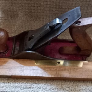May include: A wooden hand plane with a red body and a metal blade. The plane is sitting on a wooden ruler with markings in inches.