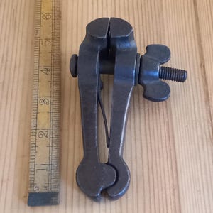 May include: A black metal hand vice with a wing nut and a spring. The vice is about 6 inches long and is sitting on a wooden surface.