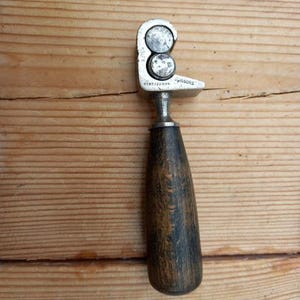 May include: A vintage metal and wood tool with a wooden handle and a metal head with two circular holes. The metal head is marked with the text "3/16" and "PISSORS".