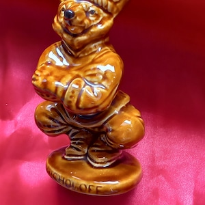 Könnte beinhalten: Eine braune Keramikfigur eines Bären, der einen Hut trägt und mit verschränkten Armen sitzt. Die Figur steht auf einem runden Sockel mit der Aufschrift "Nicholas Off".