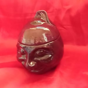 Könnte beinhalten: Eine schwarze Keramikdose mit einem Deckel in Form eines Gesichts. Die Dose hat eine strukturierte Oberfläche und der Deckel hat einen kleinen, runden Knopf oben.