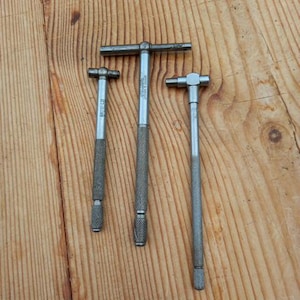 May include: Three silver-coloured metal measuring tools with a T-shaped handle and a graduated scale. The tools are laid out on a wooden surface.