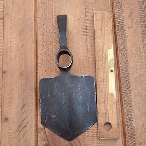 May include: A dark metal garden hoe with a flat, rectangular blade and a circular opening for a handle. A wooden ruler, marked in inches, is positioned next to the hoe. The hoe's blade is approximately 4 inches wide.