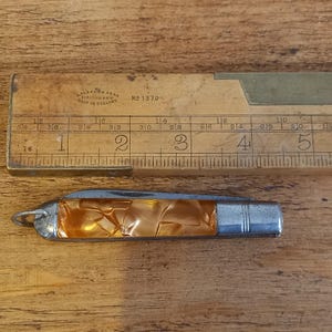 Könnte beinhalten: Ein Vintage-Taschenmesser mit einem perlmuttartigen orangefarbenen Griff und einem silberfarbenen Metallende. Ein Holzlineal mit Zentimetermarkierungen und dem Text "J. Rabone & Sons, Made in England" liegt neben dem Messer.