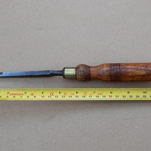 May include: A wooden handled chisel with a metal blade and brass ferrule. The handle is brown with a darker wood grain and has a brass band around it. The blade is black and has a slight curve to it. The chisel is about 18 inches long.
