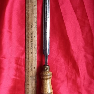 May include: A wooden handled chisel with a metal blade, measuring approximately 9 inches in length. The chisel is resting on a red fabric surface next to a wooden ruler.