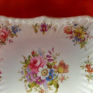Pode incluir: Um prato quadrado de servir de porcelana branca com uma borda ondulada e uma borda dourada. O prato é decorado com um padrão floral em tons de rosa, vermelho, amarelo, azul e verde.