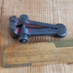 May include: A gray metal hand clamp with a red painted handle. The clamp is about 3 inches long and is sitting on a wooden surface next to a ruler.