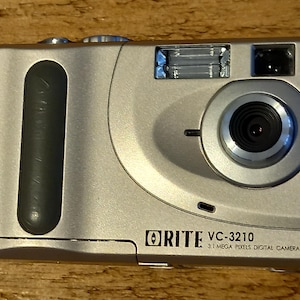 Op de afbeelding: Een zilveren digitale camera met een zwarte lens en een zwarte grip. De camera heeft de tekst "ORITE VC-3210 3.1 MEGA PIXELS DIGITAL CAMERA" aan de voorkant.