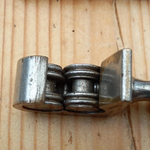May include: Close-up of a silver metal chain breaker tool with a handle and a set of rollers. The tool is designed to break and connect bicycle chains.