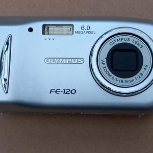 May include: A silver Olympus FE-120 digital camera with a 6.0 megapixel lens. The camera has a silver lens ring with the text "Olympus Lens AF Zoom 6.3-18.9mm 1:2.8-4.9".