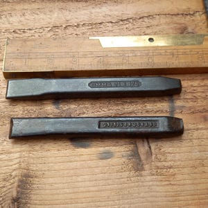 Op de afbeelding: Twee donkere metalen beitels met gestempelde tekst, één met de tekst "OMMA 21 x 175" en de andere "21/175 BRUONAOD". Een houten liniaal met messing accenten staat op de achtergrond. De gereedschappen liggen op een houten oppervlak.