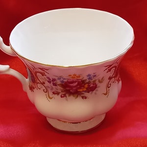 Puede incluir: Una taza de té blanca con un patrón floral en tonos de rosa, rojo y morado. La taza tiene un borde dorado y un asa delicada.