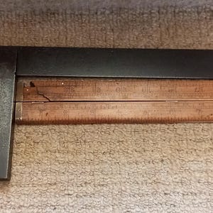 Op de afbeelding: Een zwarte metalen winkelhaak met een houten liniaal eraan bevestigd. De liniaal heeft markeringen in inches en is genummerd van 1 tot 24.