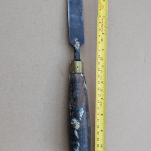 Puede incluir: Un cincel vintage con mango de madera, una virola de latón y una hoja de metal negro. El cincel mide aproximadamente 18 cm de largo.