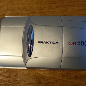 May include: A silver Praktica CM 500 camera with a silver lens cap.